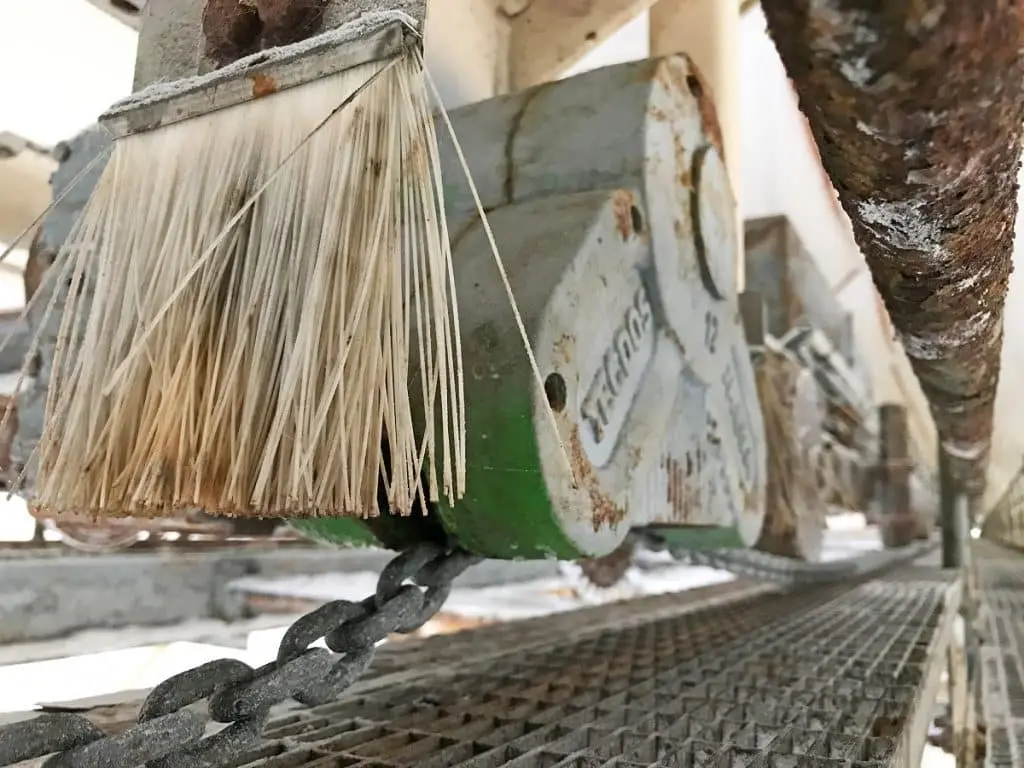 Kettenförderer in Nahaufnahme. Bürste vor dem Förderer ist zu sehen. Rechts auch ein völlig verrostetes Metallrohr.