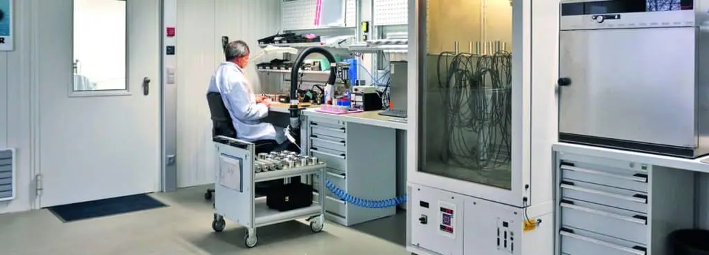 An einem elektronischen Arbeitsplatz sitzt ein Mann mit Sensoren auf einem Rollwagen neben sich. Rechts von ihm Geräte aus der Sensortechnik.