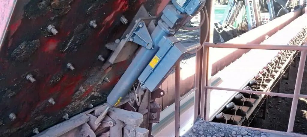 Industriehalle: Im Hintergrund ein helles Förderband, die Rollen sind gut zu sehen. Im Vordergrund eine hydraulische Anlage, die eine Platte zu bewegen scheint.
