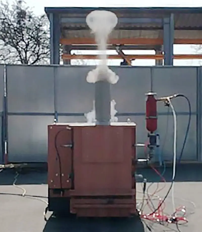 Versuchsanordnung. rötlicher Ksten mit Drähten und Leitungen verbunden und rechts einer Druckluftkartusche. Oben auf ein Kaminrohr in Stahl, aus dem Dampf einer Explosion austritt.