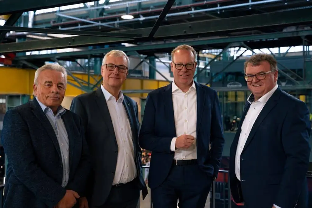 Vier Herren stehen in einer Industriehalle an einer erhöhten Position. Alle haben ein helles Hemd an und einen dunklen Anzug.
