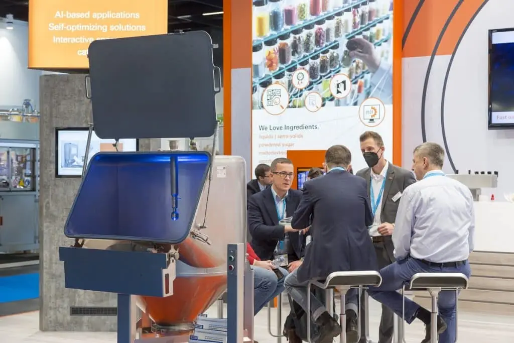 Mehrere Männer in Gespräch, teils sitzend auf Hochstühlen, teils stehend am Messestand der AZO auf der Anuga FoodTec 2022 in Köln. Im Hintergrund ein Foto mit Schüttgütern in Gläsern, die in einem Regal stehen. Eine Hand greift nach einem Glas.