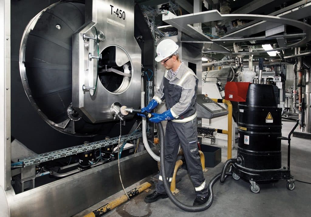 In einer Industriehalle steht ein Mitarbeiter mit einem weißen Helm und saugt eine Maschine.