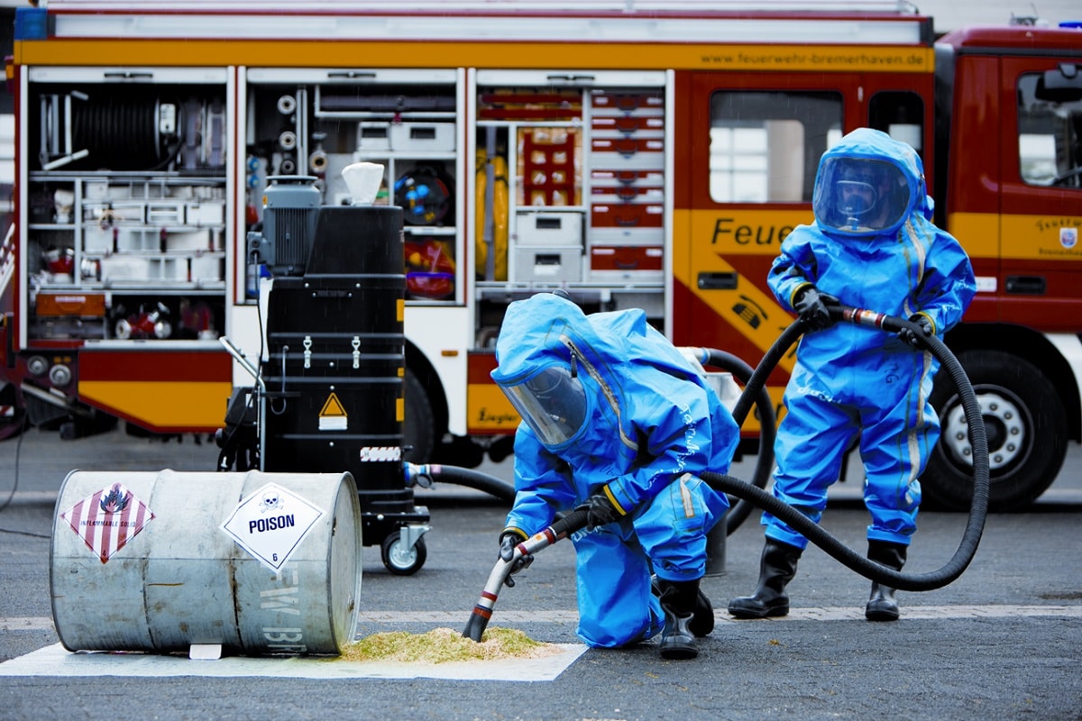 Männer der Feuerwehr in blauen Schutzanzügen mit mehrfacher Maskierung saugen giftiges Material vor einer Tonne vom Boden.