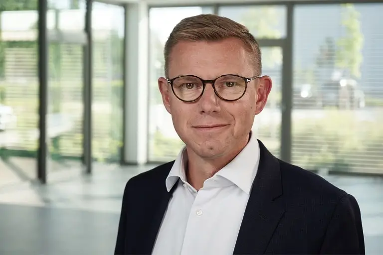 Uwe Vogt, mit weißem Hemd und schwarzen Jackett in einem Bürogebäude, mit verschwommenem Blick in den umliegenden Garten.