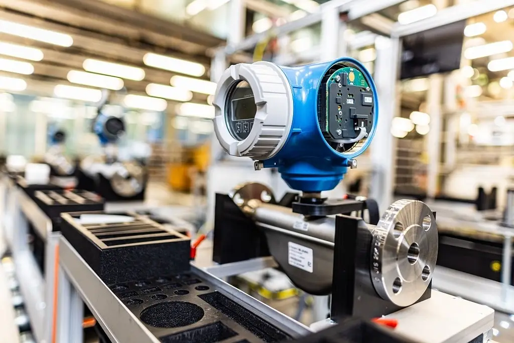 Sensor im Fertigungsprozess. Blaues Gehäuse mit weißem Rand um das Display. Die rechte Seite des Senors ist offen und man sieht die Platine. Der Sensor ist an einem Fertigungstisch an einem Rohr festgemacht.