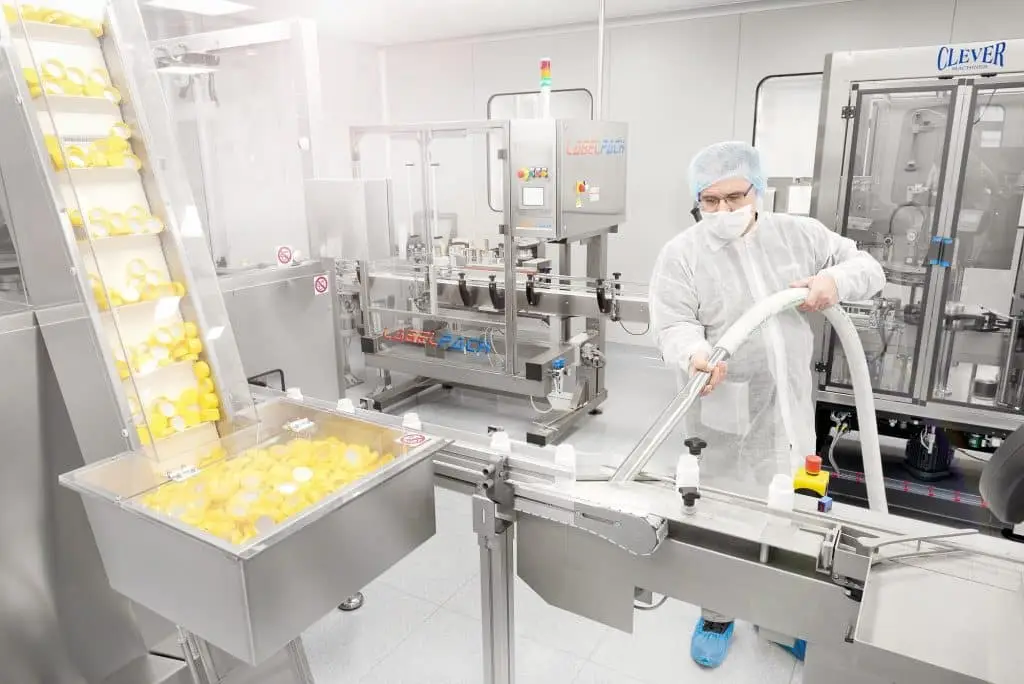 In einer Industriehalle stehen einige Produktionsmaschinen. Einen in weiß gekleideter Mitarbeiter mit blauem Plastikschutz über den Schuhen saugt eine Rinne aus.