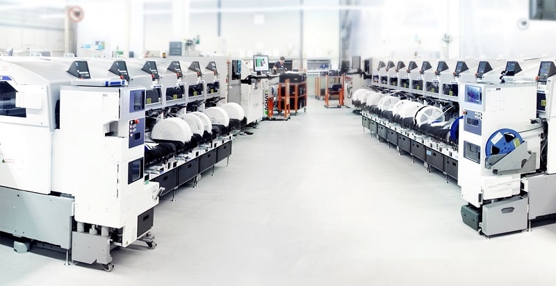 Maschinenraum mit weißen Geräten, die rechts und links aufeinander zulaufend gegenüberstehen. Im Hintergrund Arbeitsplätze mit Bildschirmen.