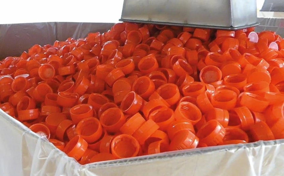 Rote Kunsstoff Plastikdelckel werden über einen Stutzen in einen Metallcontainer mit ausgelegter Folie gefüllt.