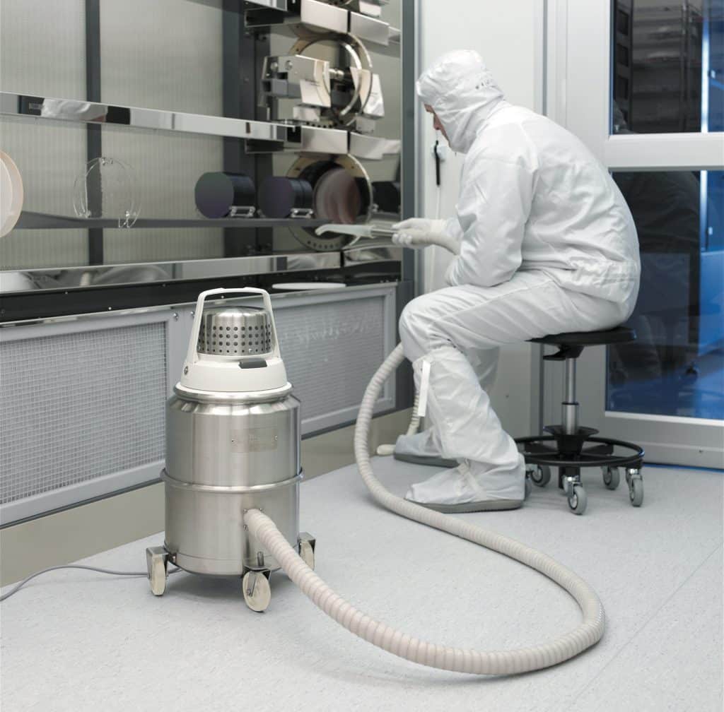 Ein komplett in weiß gekleideter Mitarbeiter saugt in einem Reinraum mit einer Plastiktülle ein Produktionsgerät aus.