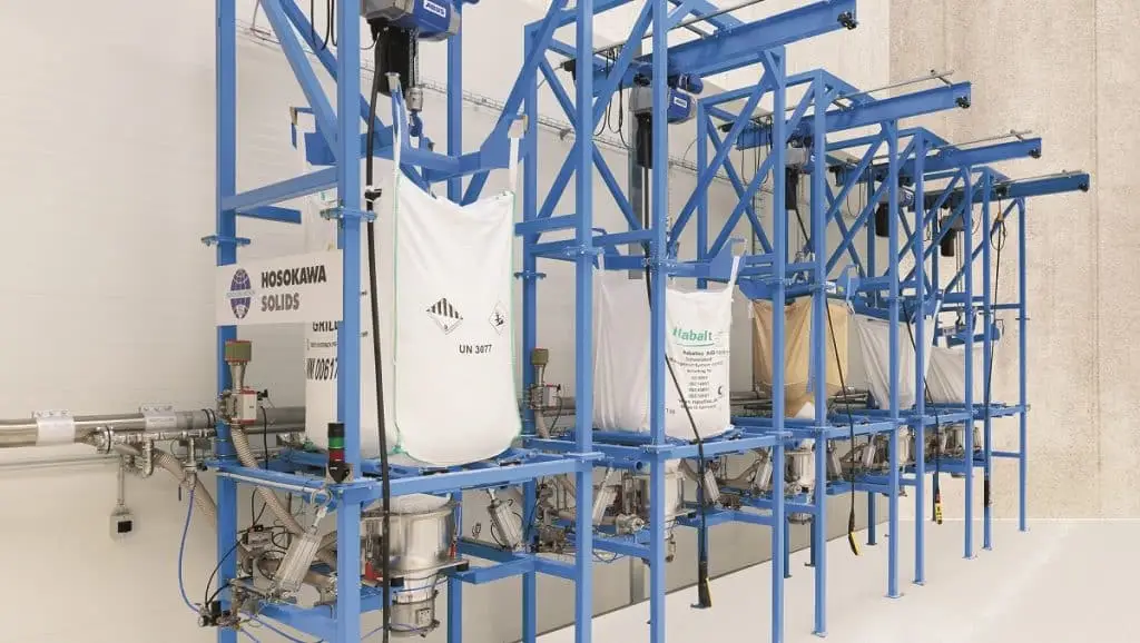 Big-Bag Entleerer in einer Industriehalle. Vorne links ein Schild Hosokawa Solids mit Logo, dann ein weißer Sack in einer Stahlkonstruktion, die blau gestrichen ist. Darunter eine Entleereinheit. Es folgen vier weitere Gestelle mit unterschiedlichen Big-Bags.