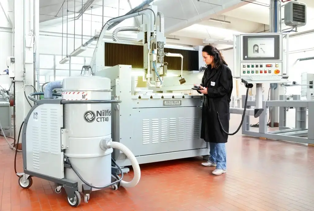 In einem Industriegebäude mit sauberen roten Fliesen steht eine Frau neben einem Sicherheitssauger vor einer Produktionsmaschine.