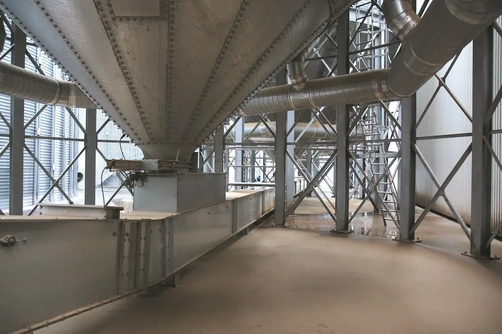 Untere Ende eines Silos mit einem Austragstrichter. Darunter ein quadratisches Metallrohr, daneben und im Hintergrund Stahlträger und viele Rohre.