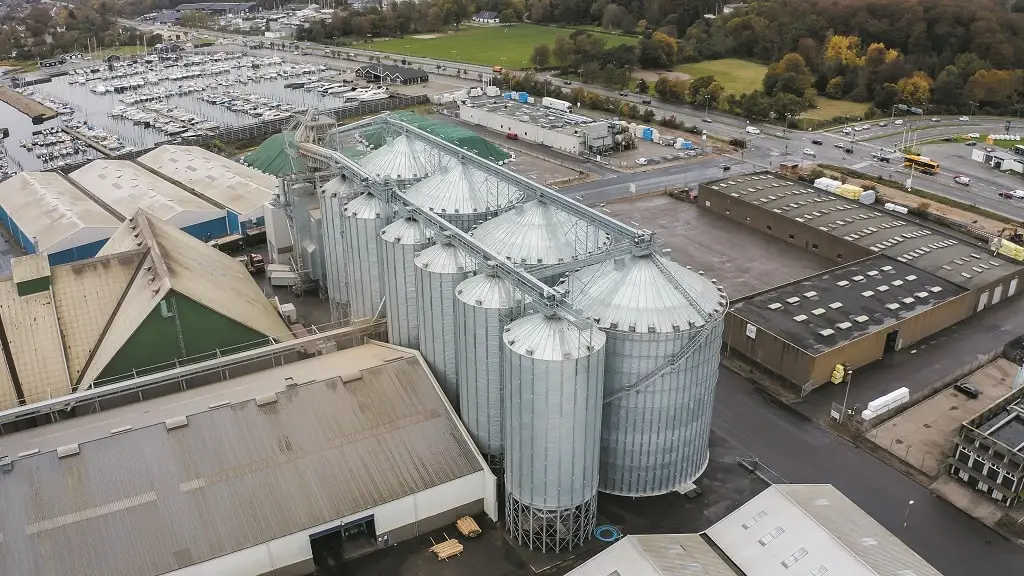 Silos an einem Segelhafen. Links Industriehallen, rechts daneben 4 große und 6 kleinere Silos, daneben ein großer, leerer Platz und andere Industriehallen, Straße und im Hintergrund der Hafen