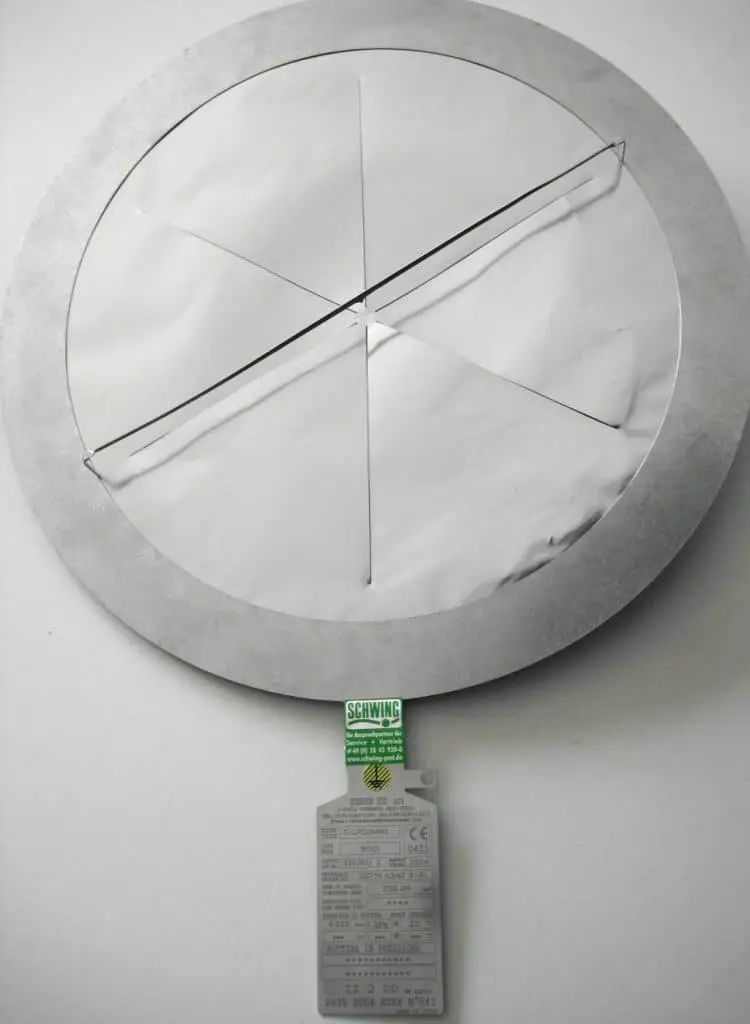 Metallring, in dem eine Berstscheibe eingebaut ist. Sie hat drei Sollbruchstellenrisse wie Pizzastücke einer Pizza. Darüber liegt ein Draht, der am inneren Ende des Rings horizontal über die Berstscheibe führt. Unten ist das Typenschild.