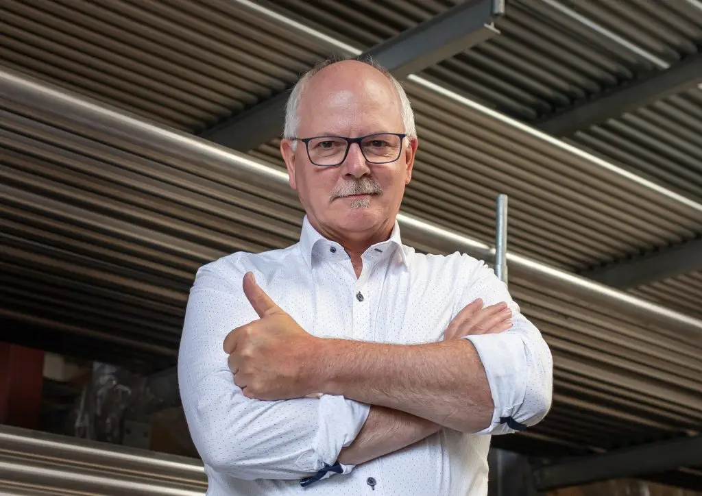 Hubertus Wessjohann steht mit überkreuzten Armen in einer Produktionshalle. Er hält den rechten Daumen hoch. Die Aufnahme ist leicht von unten gemacht.