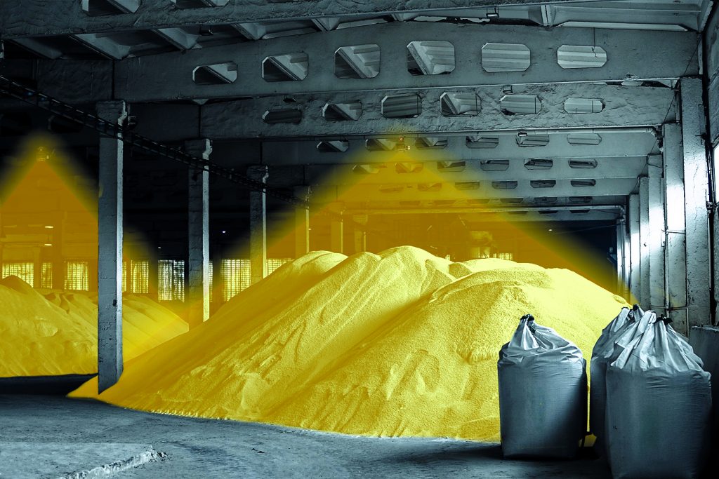 Dunkle Industriehalle mit Schüttgut-Halden, die durch einen Lichtstrahl gelb angestrahlt werden. Der Lichtstrahl ist von der Decke über die Halden grafisch sichtbar gemacht. Rechts davor stehen drei schwarze Säcke.