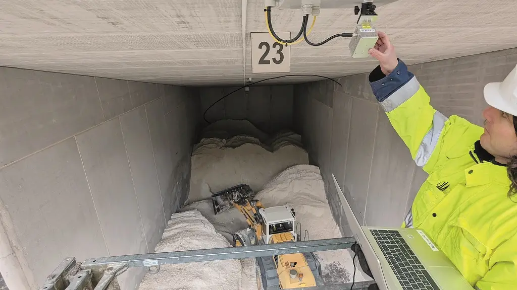 Vorn rechts ein Mitarbeiter in gelber Jacke, der eine Kamera an der Decke eines Schüttgutbunkers einrichtet. Unten im Bunker Schüttguthaufen, die von einem Bagger bewegt werden.