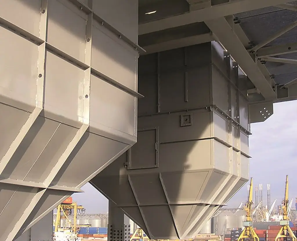 Blick von unten auf ein Silo mit einem Wägebereich. Im Hintergrund sind große Kräne und entweder eine Hafen oder Industrie-Anlagen Umgebung sichtbar.