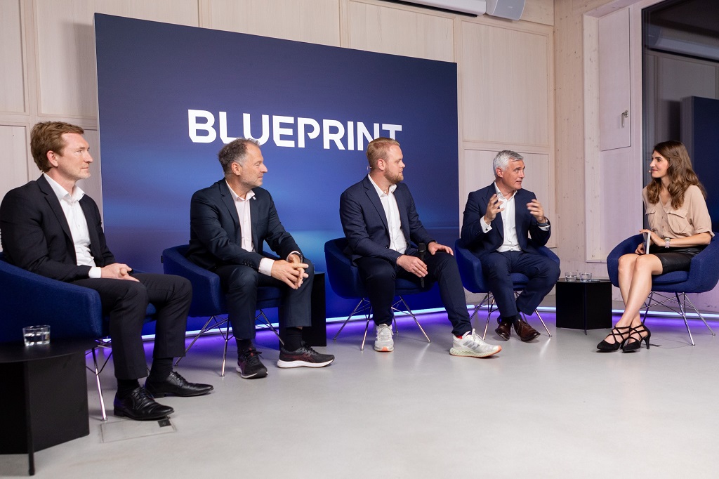 Vier Männer und eine Frau sitzen vor einer Werbewand auf der Blueprint steht und stellen sich in einer Diskussion dem Publikum.