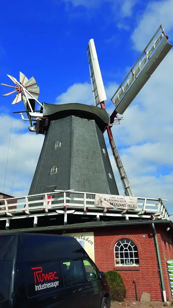 Windmühle mit schwarzen Schiefer verkleidet. Davor ein tieferes Haus mit roten Backsteine und einem Ruwac Transporter.