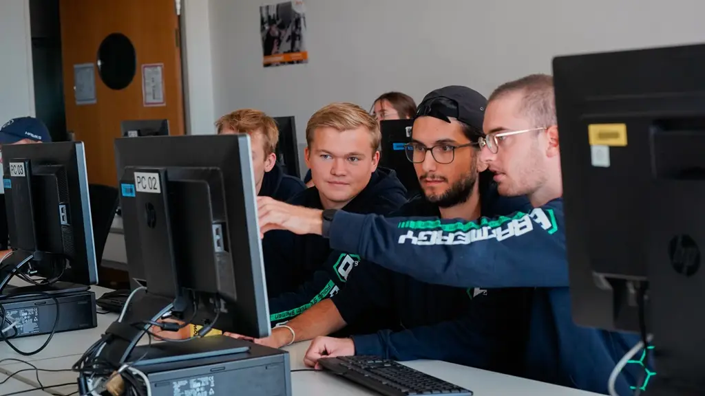 Drei junge Männer schauen in einer Reihe von PC auf einen Bildschirm und scheinen etwas zu diskutieren.