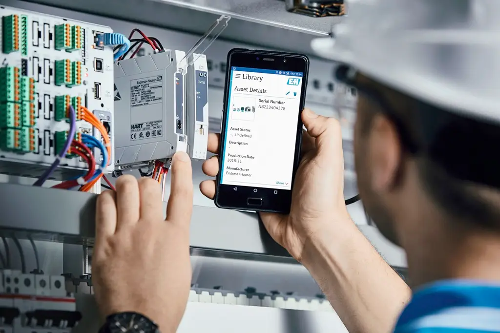 Mitarbeiter mit weißem Helm vor Schaltkasten und mobiler App der EH in der rechten Hand