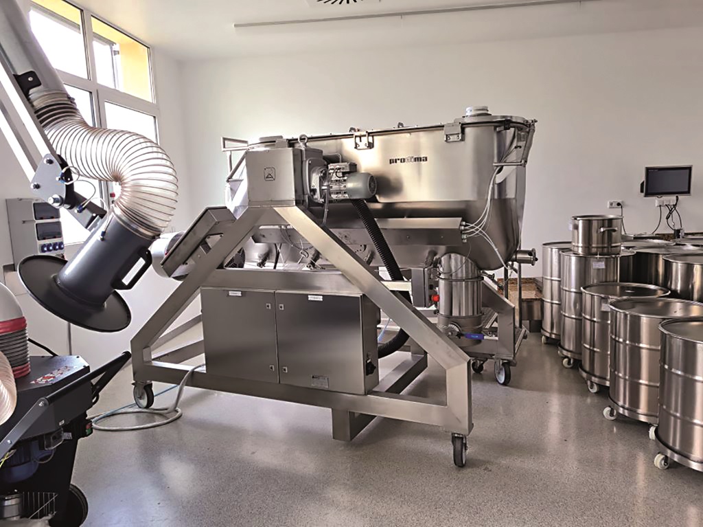 Mischer für Nahrungsergänzungsmittel in einer Industriehalle. Rechts stehen Metalltonnen auf Rädern und links der Industriemischer, auch auf Rollen.