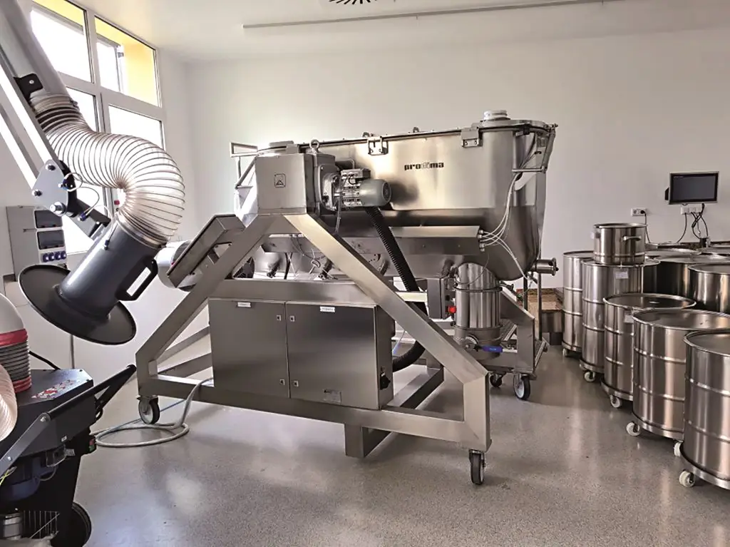 Mischer für Nahrungsergänzungsmittel in einer Industriehalle. Rechts stehen Metalltonnen auf Rädern und links der Industriemischer, auch auf Rollen.