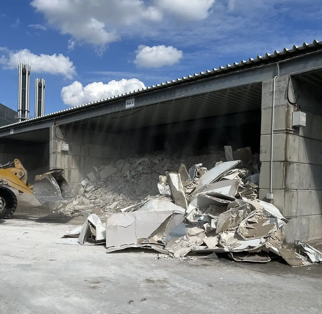 Staubbindeanlage, die vorn, oberhalb des überdachten Lagers Wasser sprüht, bindet den Staub vor einem Bauschuttbunker, der mit einem Bagger entleert wird.