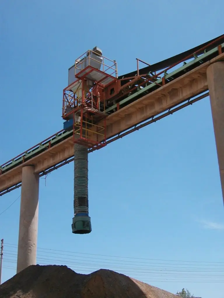 Außengelände, blauer Himmel und unter einer Art Brücke ist eine Schüttguthalde. Oben ein Telescoplader, dessen Schlauch über der Halde steht.