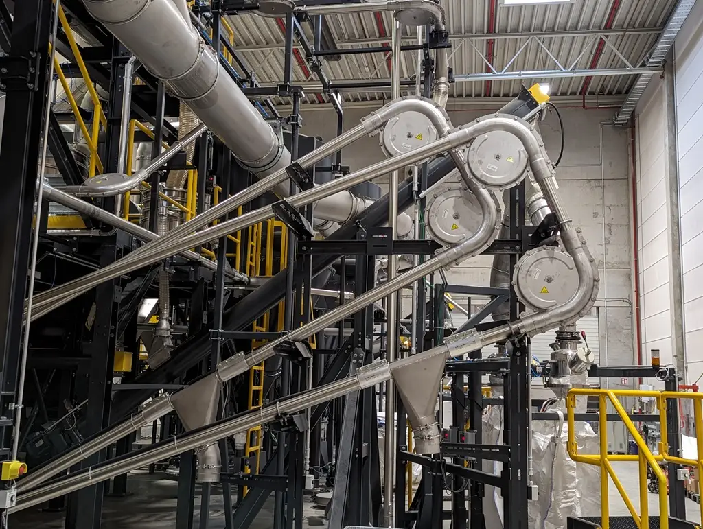 Rohr-Seilzug-Förderer in einer Industriehalle. Man sieht die Rohre und eine Runde Verteiltechnik an den Abrundungen der Rohrführung.