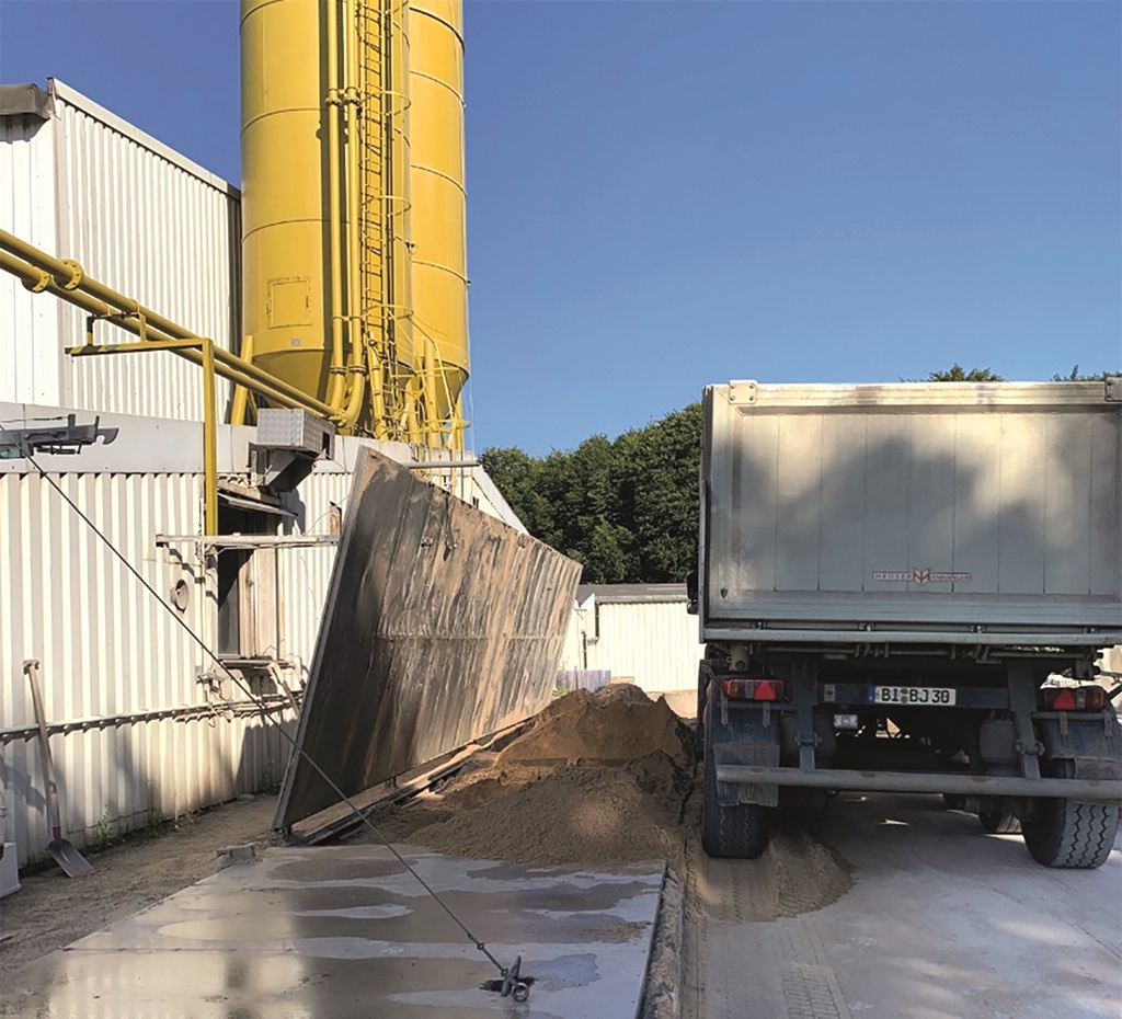 Links ein Industriegebäude und Silo in der Mitte ein Bunker für Gestein, dessen Deckel geöffnet ist, Rechts ein Lastwagen.