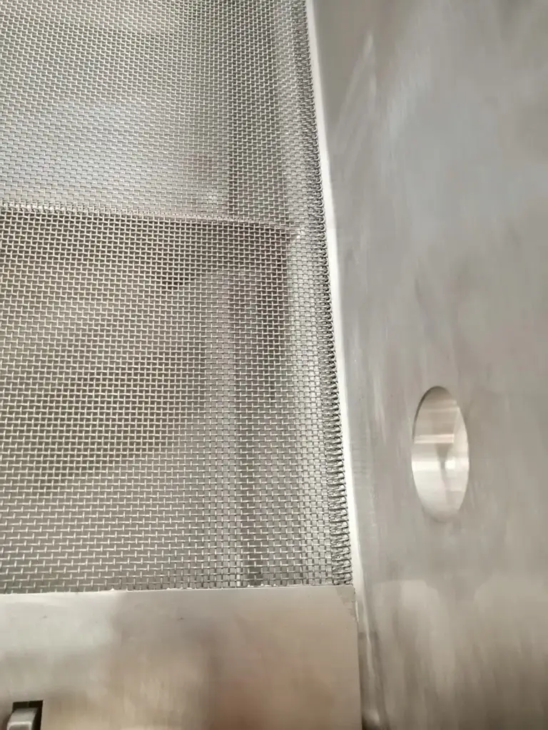 Links ein Sieb, dessen Rand vorne mit einem Blech abgedeckt ist. Rechts auf der glatten Seitenbegrenzung eine CIP Düsenhülle