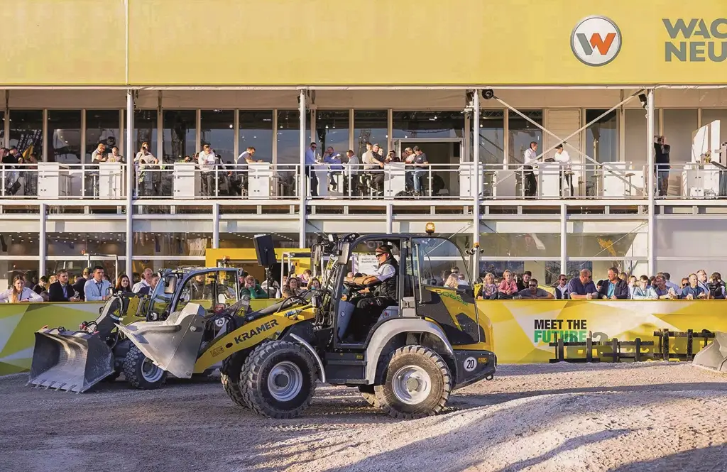 Außengelände der bauma 2024. Im Vordergrund ein Kramer Bagger auf sandigem Untergrund. Dahinter eine Trennbande mit Zuschauern. Im Hintergrund ein Messegebäude.