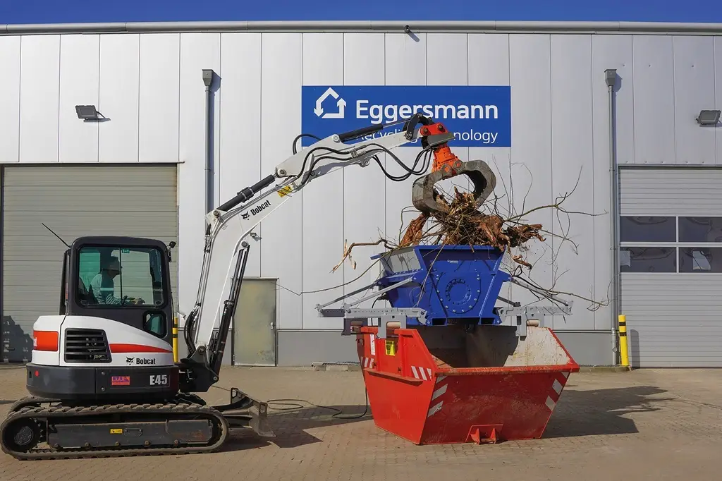 Rechts ein Radlader, der mit einem kleinen Kran den Zerkleiner mit Holzmaterial füllt. Unter dem Zerkleinerer steht ein handelsüblicher Container
