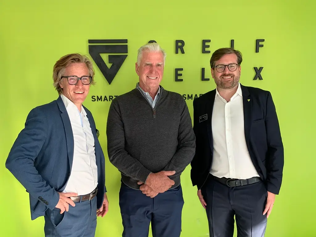 Tobias Elsner (v.l., Greif-Velox), John E. Thompson (Trans Automation Technologies) und Sebastian Pohl (Greif-Velox) vor einer grünen Wand auf der Greif Velox steht.
