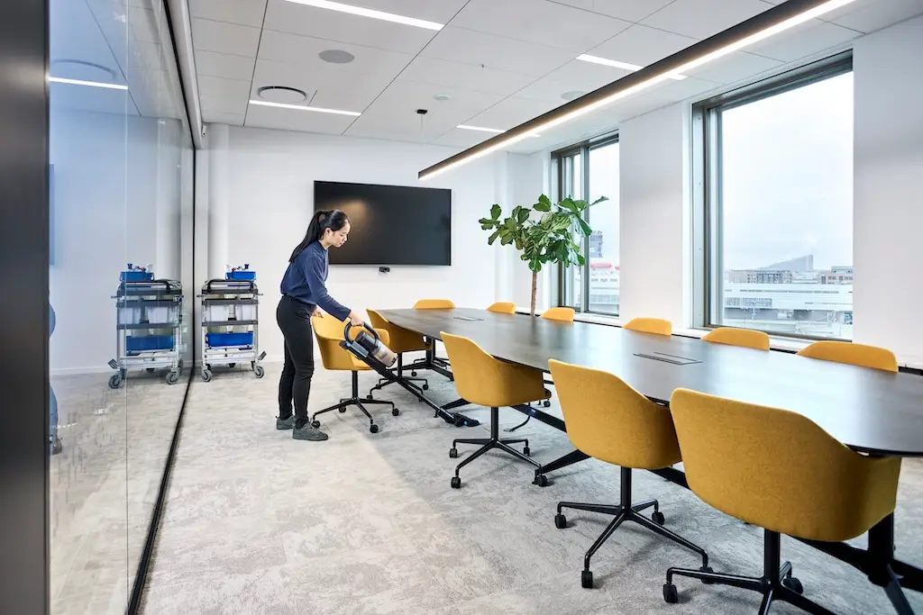 Konferenzraum mit einer langen Tafel. Der Raum wird von einer Putzkraft mit einem Staubsauger gesaugt