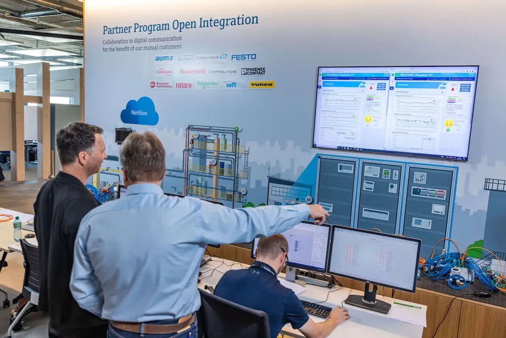 Büro mit mehreren Bildschirmen, vor denen drei Männer stehen bzw. sitzen.