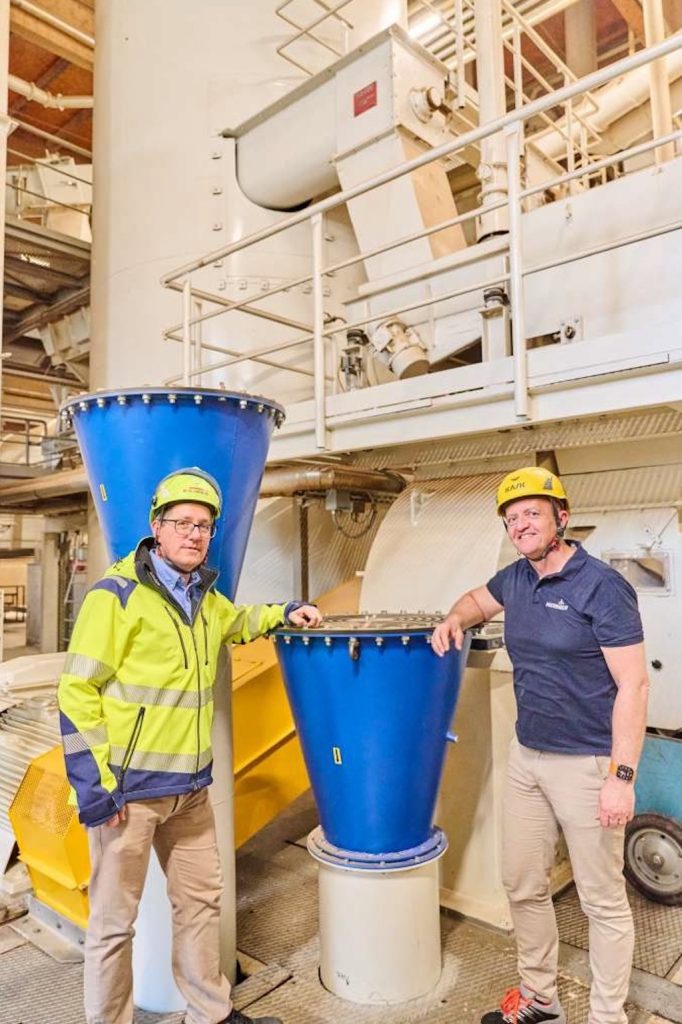 Zwei Mäner stehen in Arbeitskleidungvor einer groen Holzverarbeitungsmaschiene