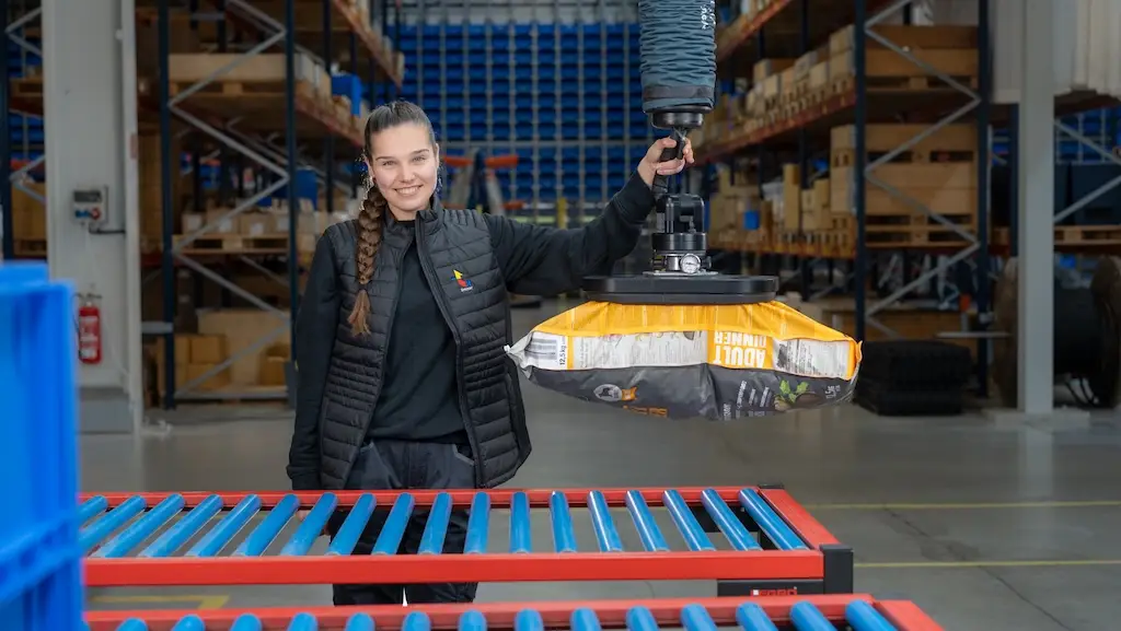 Lagerhalle im Hintergrund. Im Vordergrund eine Frau die mit Hilfe einer hebehilfe einen Sack von einem Förderband hebt