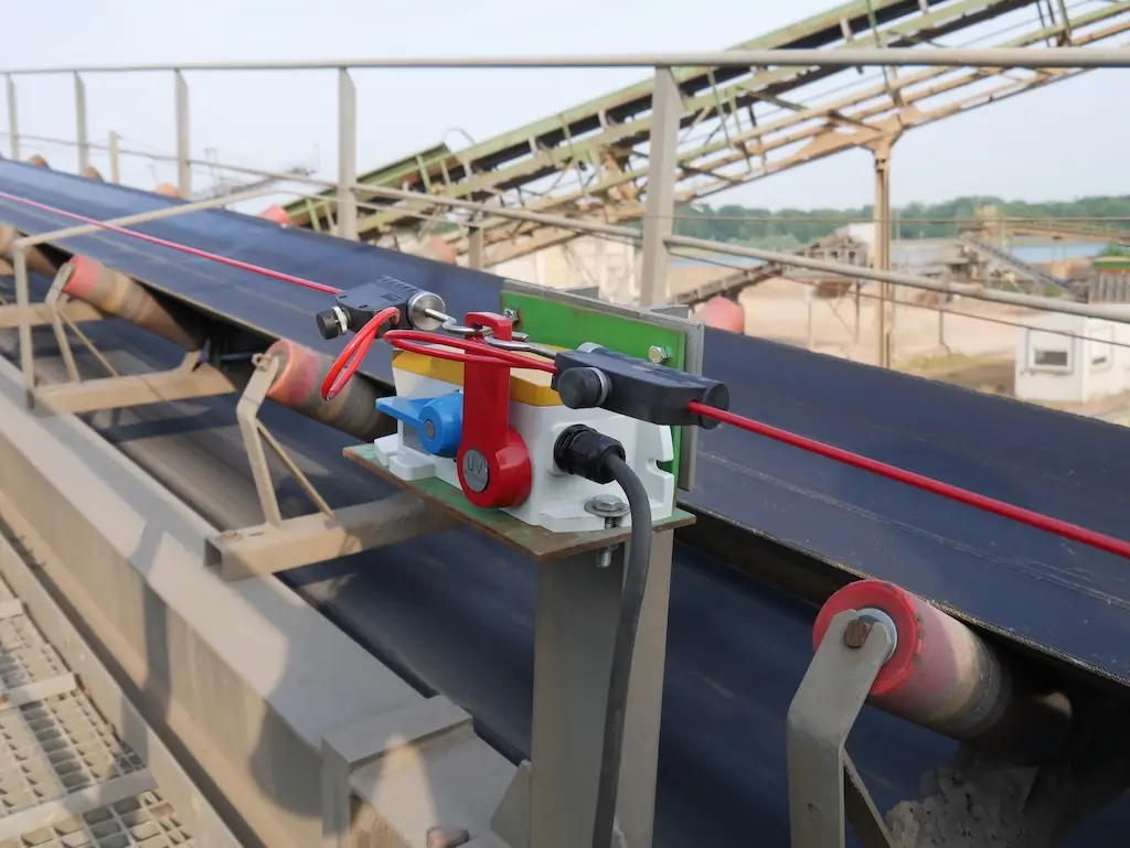 Blick auf ein Förderband. Im Vordergrund ein Seilzug-Notschalter. Im Hintergrund eine Kiesgrube.