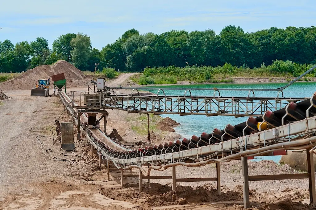 Vor einem See ist ein Förderband, an dessen Ende ein Bagger steht und ein Trichter zum Dosieren auf das Förderband.