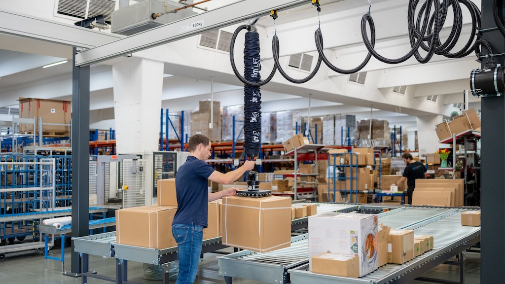 Lagerhalle mit Kartons. Im vordergrund einnmitarbeiter der eien Karton mit einer hebehilfe bewegt