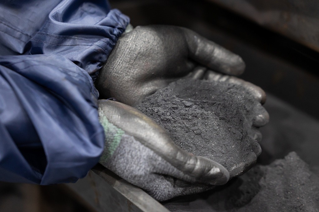 Behandschuhte Hände halten eine Menge feines graues Pulvermaterial in einem industriellen Umfeld.