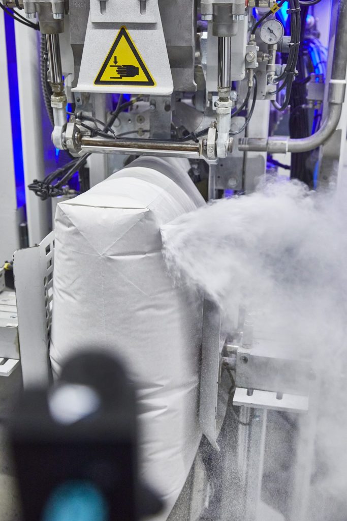 Industrielle Füllung eines Sacks mit pulvrigem Material. Der Sack ist oben rechts undicht und das Material sprüht heraus