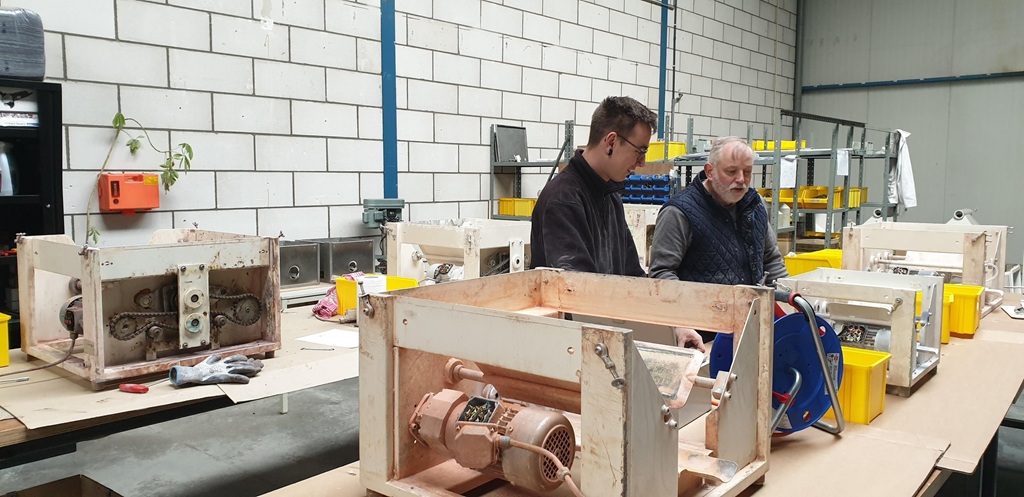 Werkstatt mit mehreren geöffneten Industrie‑ und Maschinengehäusen, an denen zwei Personen technische Reparaturen und Wartungsarbeiten durchführen. Auf den Tischen stehen Bauteile wie Motoren, Zahnräder und elektrische Komponenten. Im Hintergrund befindet sich ein Regal mit gelben Behältern und Werkzeugen, typisch für eine industrielle Instandhaltungs‑ oder Refurbishment‑Umgebung.