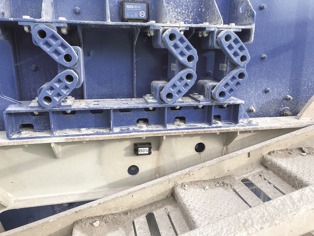 Detailaufnahme eines blauen Industriebauteils mit mehreren rechteckigen, gitterförmigen ROSTA-Elementen, montiert an einer Metallstruktur einer Siebanlage. Im unteren Bereich ist ein kleiner Sensor angebracht; die Umgebung ist staubig und zeigt typische Gebrauchsspuren aus dem Schüttgutbetrieb.