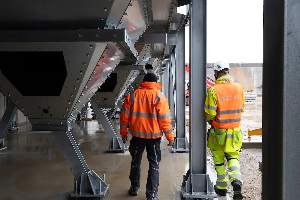 Bauarbeiter in Warnschutzkleidung und Helmen inspizieren eine Stahlkonstruktion bzw. Förderanlage für Schüttgut in einer modernen Industrieanlage.
