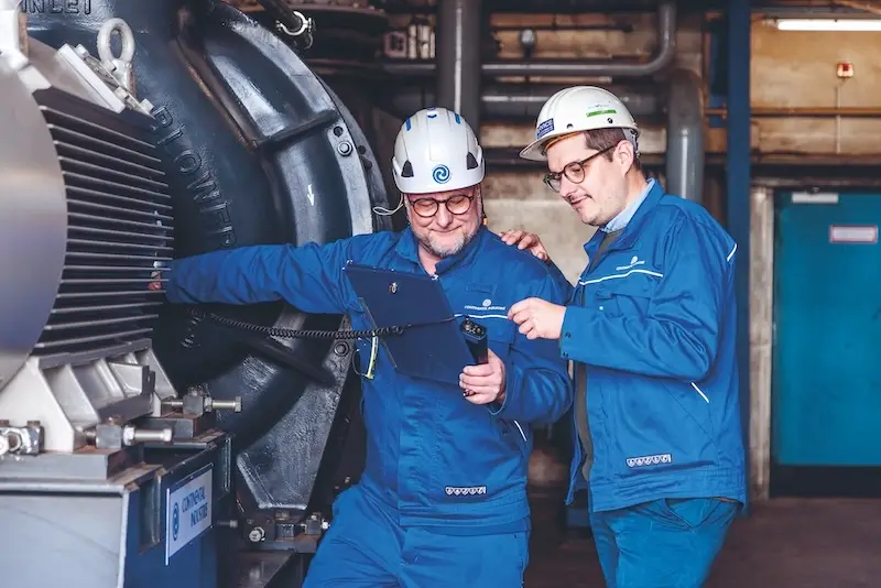 Zwei Continental Industrie Techniker in blauer Arbeitskleidung und Helmen begutachten gemeinsam Daten auf einem Tablet vor einem großen Industriegebläse